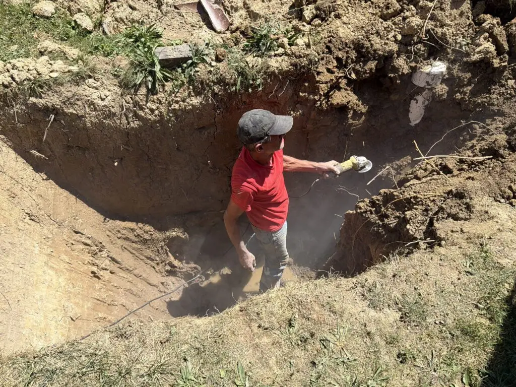 Technician in a deep trench cutting a white PVC pipe during underground repair