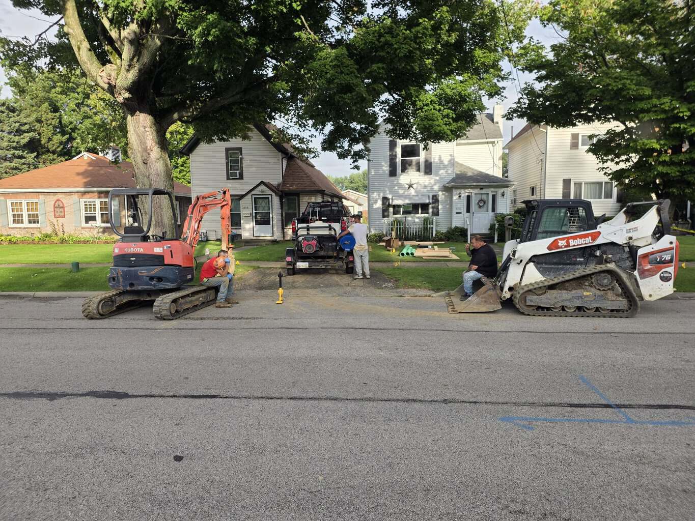 Mini-excavator, track loader, and crew staged for underground plumbing work on a residential street
