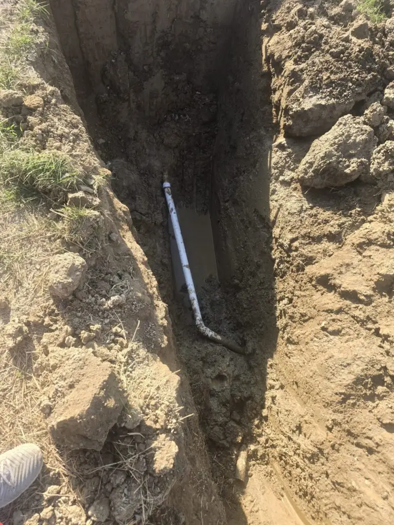 White PVC pipe exposed at the bottom of a deep muddy excavation trench