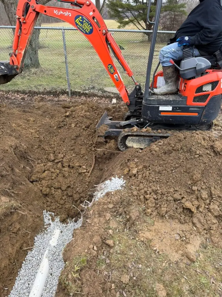 Orange mini-excavator beside a trench with PVC pipe in gravel bedding